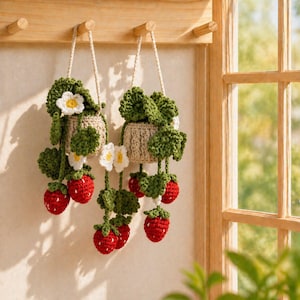 May include: Two hanging crochet planters made of beige yarn, each containing green crocheted leaves, white flowers with yellow centers, and dangling red strawberries with small white embroidered seeds, suspended by matching beige yarn loops.