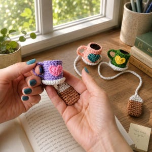 May include: Three miniature crochet coffee cup bookmarks, each featuring a small heart on the side and a dangling brown tea bag. One cup is purple with a pink heart, one is pink with a teal heart, and one is green with a yellow heart.