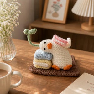 May include: A crocheted white duck figurine with an orange beak and feet, wearing a pink hat. The duck rests on a brown square base next to a stack of light blue and yellow crocheted squares, with a small green sprout extending from its head.