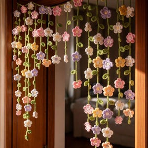 Puede incluir: Una cortina floral de ganchillo en colores pastel cuelga en una puerta. La cortina presenta múltiples hebras de flores de ganchillo en tonos rosa, amarillo, morado y blanco, con vides y hojas verdes.