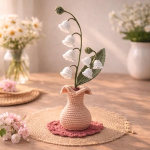 Puede incluir: Un arreglo de lirios de los valles de ganchillo en un pequeño jarrón color melocotón. Las flores hechas a mano son blancas, con tallos y hojas verdes. El jarrón se asienta sobre un tapete redondo de ganchillo rosa, con un salvamanteles de fibra natural debajo.