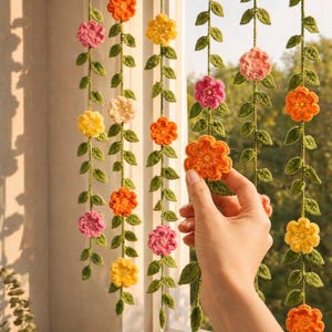 May include: Vertical hanging garlands featuring crocheted green vines with small leaves and multi-colored flowers in shades of yellow, orange, and pink. The floral strands are suspended in front of a window, showcasing intricate yarn textures.
