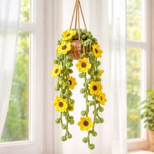 May include: A crocheted hanging plant featuring bright yellow sunflowers with dark brown centres and long, trailing green vines with small round leaves. The plant is contained in a small, textured, light brown woven basket suspended by a rope.