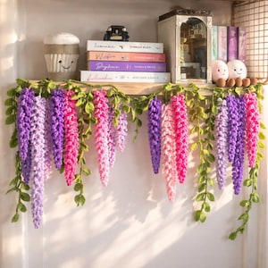 Puede incluir: Una estantería decorada con una guirnalda de glicinas de ganchillo en tonos morados y rosas, con hojas verdes. La estantería también contiene libros, una magdalena decorativa y otros objetos pequeños.