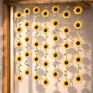 May include: Crocheted sunflower garland with yellow petals and brown centers, strung on green vines. The garland is suspended from a wooden rod, creating a decorative curtain or wall hanging. The sunflowers are evenly spaced.