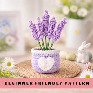 May include: A crocheted lavender plant in a purple pot with a white heart design. The lavender has purple flowers and green stems. The pot is sitting on a round, woven mat. Text at the bottom reads "BEGINNER FRIENDLY PATTERN".