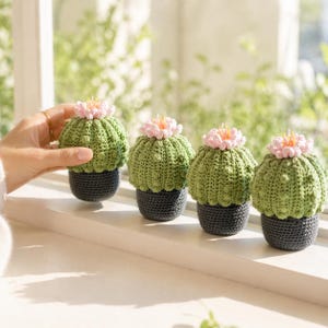 May include: Four handmade crochet cactus plants with pink flower accents sit on a white windowsill. The cacti are green with a textured pattern, and the pots are dark grey. The background shows a blurred view of green foliage.