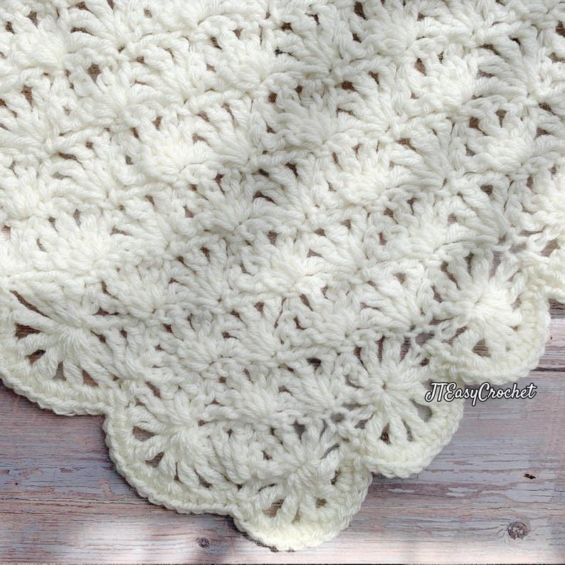 May include: Cream-colored, handmade crochet blanket with a scalloped edge. The blanket features a detailed, textured pattern of fan-like stitches. The overall design is soft and inviting, perfect for a cozy home decor item.