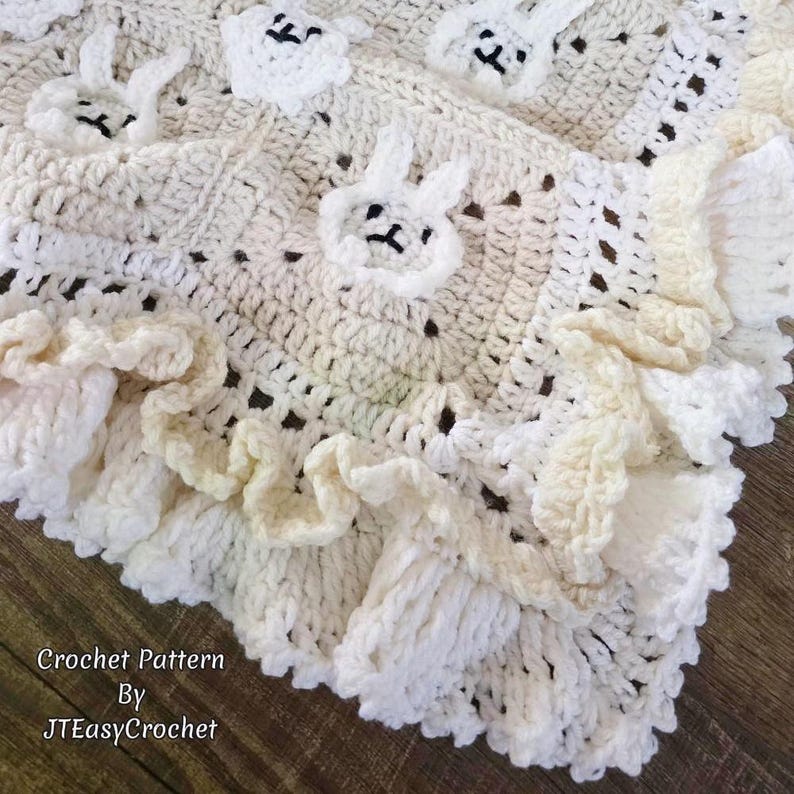 Beautiful Bunny  granny square blanket with fringed edging is laying on a brown hardwood surface. The blanket is cream with white bunnies on each square. The lettering says:  “Crochet Pattern By JTEasyCrochet” meaning that JTEasyCrochet has the best crochet patterns and fun crochet ideas.
