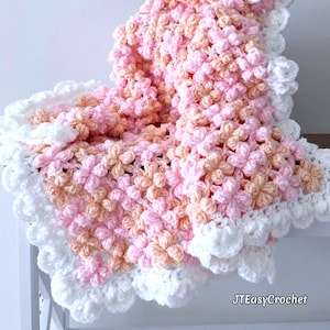 May include: A handmade crochet blanket in shades of pink, peach, and white. The blanket features a floral pattern and a scalloped white border. The text "JTEasyCrochet" is visible in the bottom right corner. This cozy blanket is perfect for a baby.