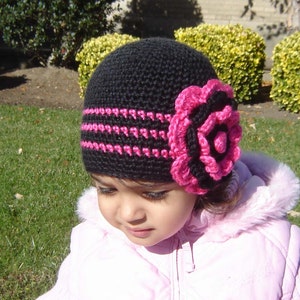 Puede incluir: Un gorro de punto negro con un adorno de flor rosa. El gorro tiene una banda negra con rayas rosas.
