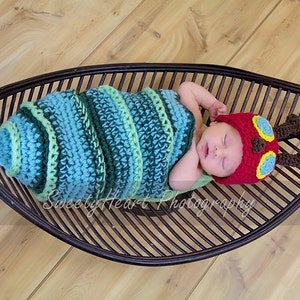 May include: A newborn baby wearing a crocheted caterpillar costume and a red hat with yellow and blue accents, lying in a brown wicker leaf-shaped basket.