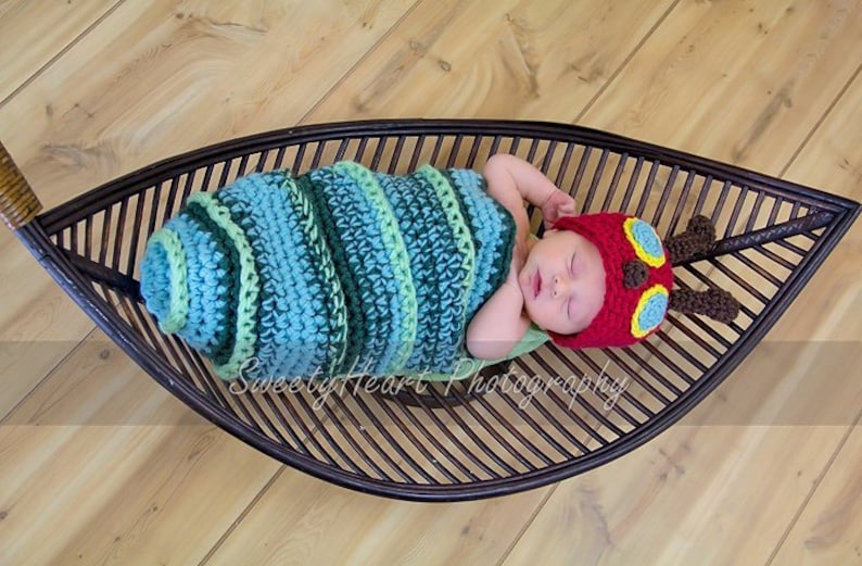 May include: A newborn baby wearing a crocheted caterpillar costume and a red hat with yellow and blue accents, lying in a brown wicker leaf-shaped basket.