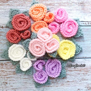 May include: A collection of handmade crochet flowers in various colors, including pink, orange, yellow, white, and purple. Each flower has a detailed spiral design, with green leaves. The flowers are arranged on a light blue wooden surface. The text "JTEasyCrochet" is visible.