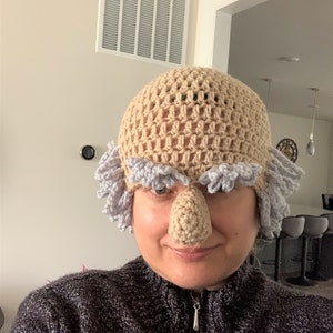 May include: A crocheted hat with a large, brown nose and gray hair. The hat is worn by a person with a gray sweater.