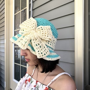 Puede incluir: Un sombrero de sol de crochet azul claro y blanco con un gran lazo de crochet blanco. El sombrero lo lleva una persona con el pelo oscuro.