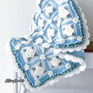 Beautiful crochet granny square blanket is hanging from a display on a light grey background. Each white granny square has a white textured gardenia flower in the center, framed with pale green leaves and light frosty blue border, so the whole blanket remind of a frozen gardenia blanket. There are also bunches of gardenia flowers in the image to highlight the likeness. The lettering logo says: " JTEasyCrochet"