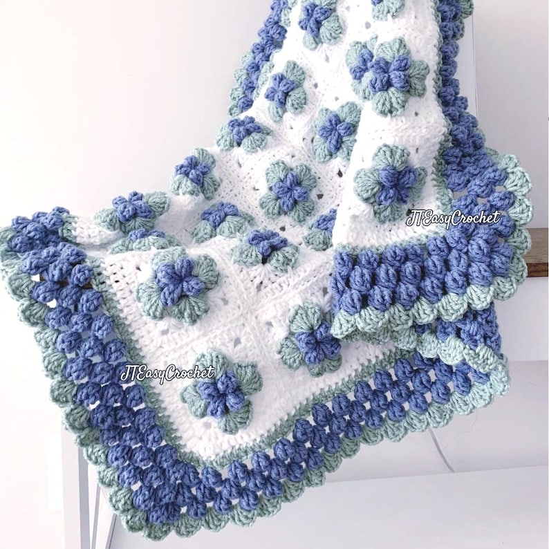Beautiful white granny square blannket is hanging from a display. In the center of each square is blueberry with green leaves. The border is made of blueberries.