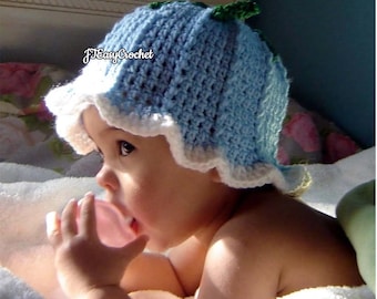May include: A baby wearing a blue and white crocheted hat with a green leaf detail. The hat has a scalloped edge. The baby is looking to the side and has a pink pacifier in their mouth.