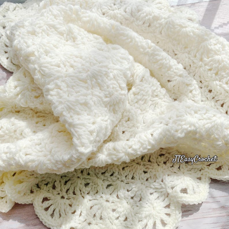 May include: Cream-colored, handmade crochet blanket with a textured stitch pattern. The blanket is folded, revealing the intricate details of the yarn and the scalloped edge. The blanket is soft and inviting, perfect for adding warmth and style to any space. The text "JTEasyCrochet" is visible.