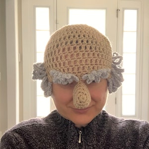 May include: A crocheted hat with a large, crocheted nose and gray hair. The hat is beige and the hair is gray.