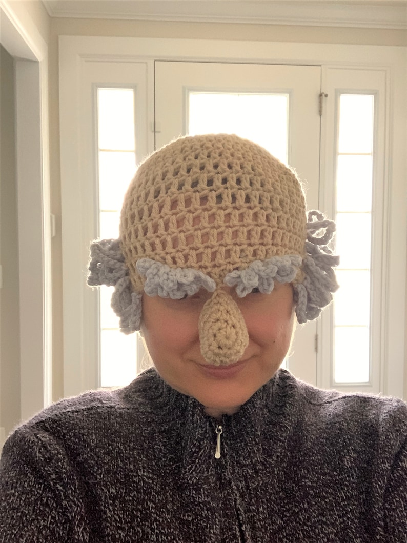 May include: A crocheted hat with a large, crocheted nose and gray hair. The hat is beige and the hair is gray.
