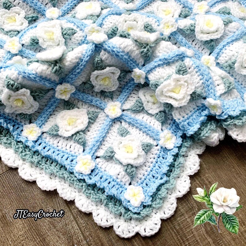 Beautiful crochet granny square blanket is tossed on the wooden floor. Each white granny square has a white textured gardenia flower in the center, framed with pale green leaves and light frosty blue border, so the whole blanket remind of a frozen gardenia blanket. There are also bunches of gardenia flowers in the image to highlight the likeness. The lettering logo says: " JTEasyCrochet"