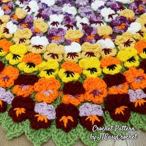 A handmade crochet blanket featuring a vibrant floral pattern. The blanket showcases a variety of colorful pansies in shades of yellow, orange, purple, and white, set against a green leaf-shaped border. The text "Crochet Pattern by JTEasyCrochet" is visible.