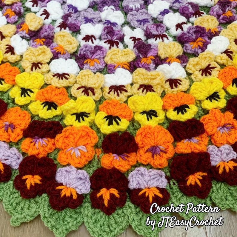 A handmade crochet blanket featuring a vibrant floral pattern. The blanket showcases a variety of colorful pansies in shades of yellow, orange, purple, and white, set against a green leaf-shaped border. The text "Crochet Pattern by JTEasyCrochet" is visible.