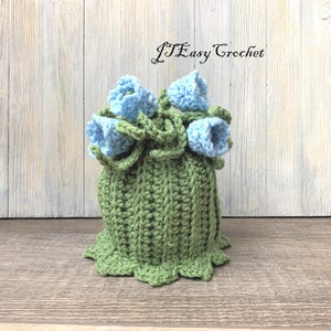 May include: A crocheted flower pot with a green base and light blue flowers. The pot has a scalloped edge and is sitting on a wooden surface. The text "J'Easy Crochet" is visible in the background.