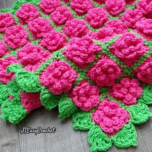 Op de afbeelding: Een handgemaakte gehaakte deken met felroze rozen en groene bladeren. De deken heeft een textuurontwerp met verhoogde bloemelementen. De tekst "JTEasyCrochet" is zichtbaar. Een gezellig en decoratief item.