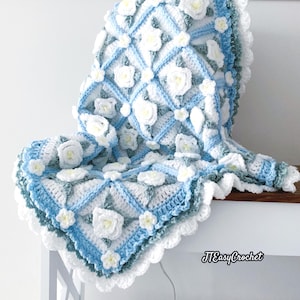 Beautiful crochet granny square blanket is hanging from a display on a light grey background. Each white granny square has a white textured gardenia flower in the center, framed with pale green leaves and light frosty blue border, so the whole blanket remind of a frozen gardenia blanket. There are also bunches of gardenia flowers in the image to highlight the likeness. The lettering logo says: " JTEasyCrochet"