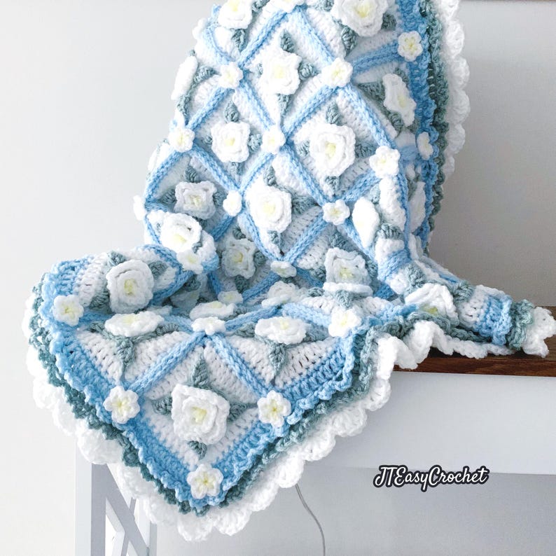 Beautiful crochet granny square blanket is hanging from a display on a light grey background. Each white granny square has a white textured gardenia flower in the center, framed with pale green leaves and light frosty blue border, so the whole blanket remind of a frozen gardenia blanket. There are also bunches of gardenia flowers in the image to highlight the likeness. The lettering logo says: " JTEasyCrochet"