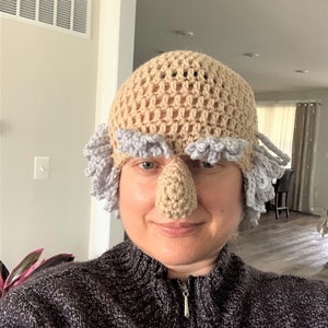 May include: A person wearing a crocheted hat with a large nose and gray hair. The hat is beige with a light purple trim.