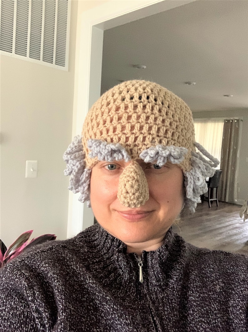 May include: A person wearing a crocheted hat with a large nose and gray hair. The hat is beige with a light purple trim.