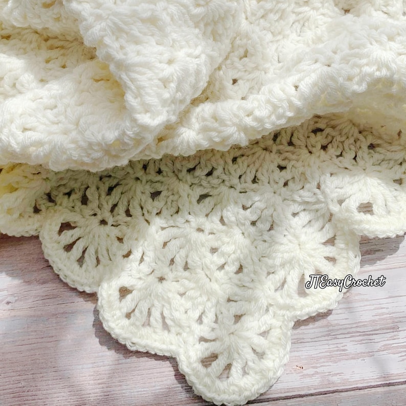 May include: Cream-colored crochet blanket with a scalloped edge. The blanket is folded, revealing the intricate stitch pattern and texture. The text "JTEasyCrochet" is visible on the edge of the blanket.
