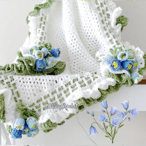 May include: A white crochet blanket with a green and white scalloped edge. The blanket is adorned with blue and yellow crochet flowers and green leaves. The text "JTEasyCrochet" is visible on the blanket.