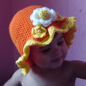 May include: A baby wearing a crocheted hat with yellow and orange flowers. The hat has a scalloped edge and a white flower with a yellow center.
