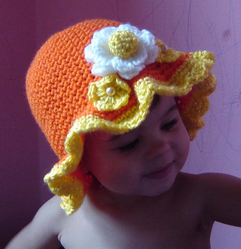 May include: A baby wearing a crocheted hat with yellow and orange flowers. The hat has a scalloped edge and a white flower with a yellow center.