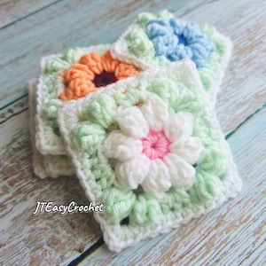 Op de afbeelding: Close-up van verschillende handgemaakte gehaakte vierkanten. Elk vierkant heeft een bloemontwerp in het midden, met kleuren als wit, roze, oranje en blauw. De vierkanten zijn geplaatst op een lichtblauwe houten ondergrond. De tekst "JTEasyCrochet" is zichtbaar.