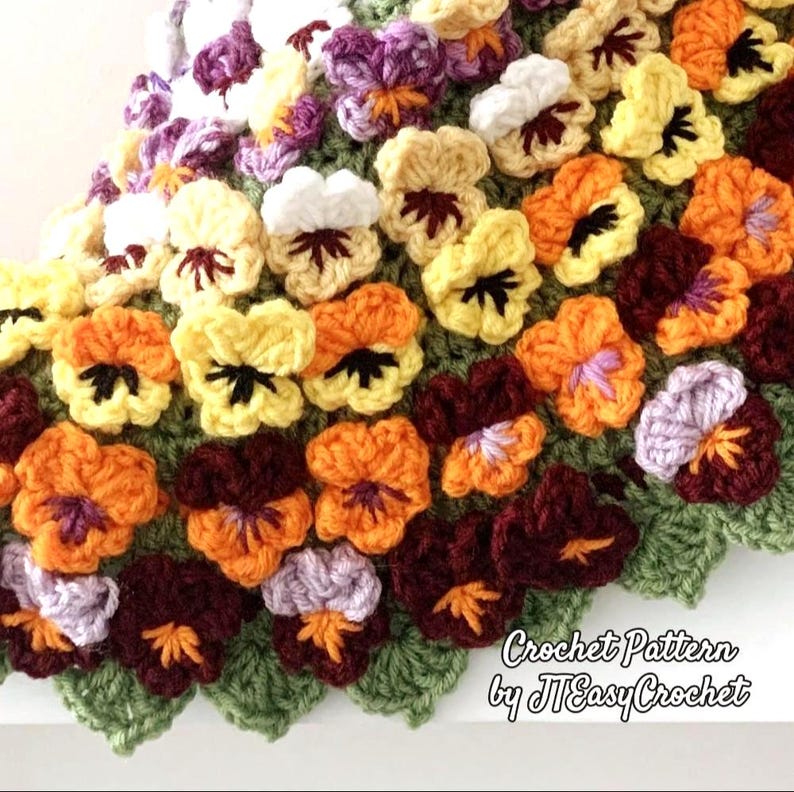 A close-up of a crocheted blanket featuring colorful pansies. The flowers are in shades of yellow, orange, purple, and burgundy, with green leaves. The text "Crochet Pattern by JTEasyCrochet" is visible.