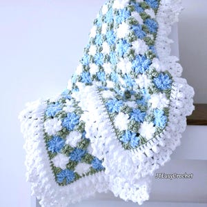 Lavishly beautiful crochet floral blanket made of cornflower motifs is hanging from a display. The blanket resembles a field of white and blue cornflowers framed with a beautiful lace edging.  The lettering says "JTEasyCrochet" meaning that JTEasyCrochet has the best crochet patterns for flower baby blankets, throws and afghans.