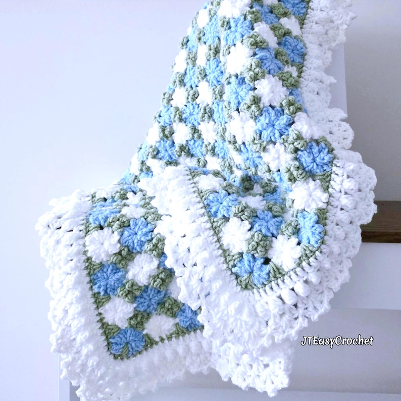 Lavishly beautiful crochet floral blanket made of cornflower motifs is hanging from a display. The blanket resembles a field of white and blue cornflowers framed with a beautiful lace edging.  The lettering says "JTEasyCrochet" meaning that JTEasyCrochet has the best crochet patterns for flower baby blankets, throws and afghans.