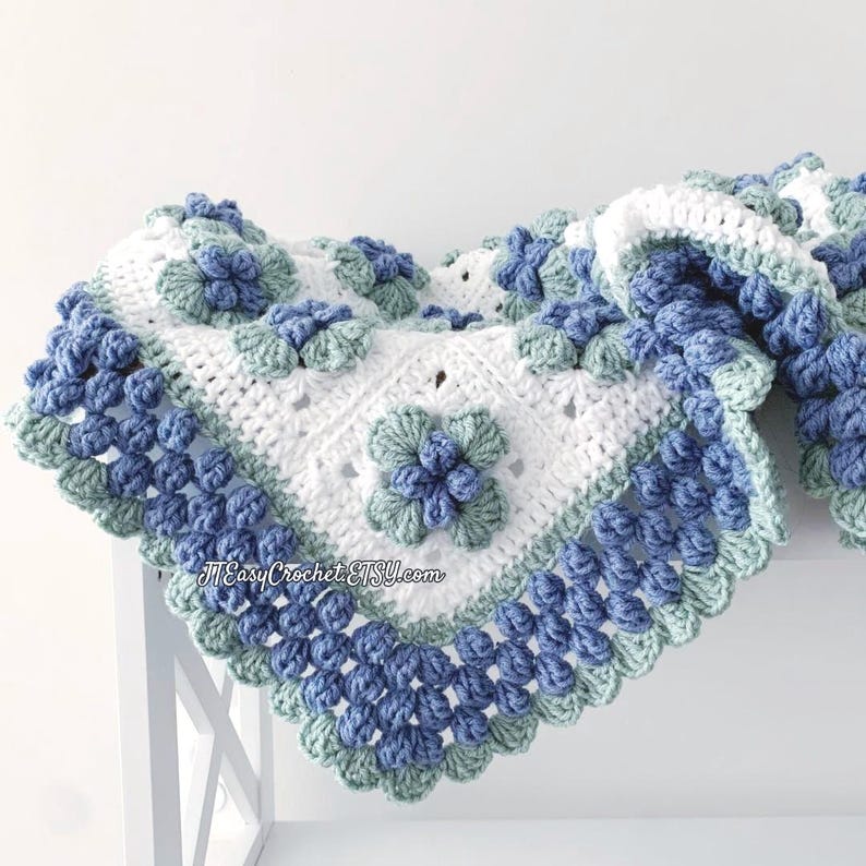 Beautiful white granny square blannket is laying on a shelf. In the center of each square is blueberry with green leaves. The border is made of blueberries. The background is light cream.