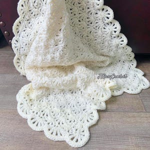 May include: A cream-colored, crocheted blanket with a scalloped edge. The blanket is folded, revealing the intricate stitch pattern. The text "JTEasyCrochet" is visible in the lower right corner. The blanket is made of soft yarn.