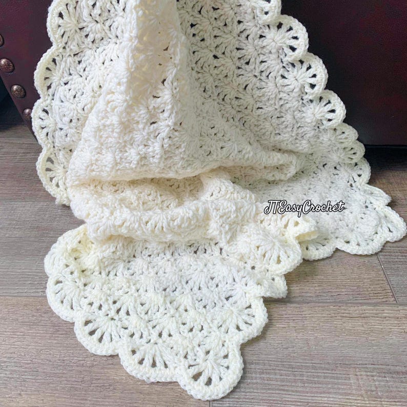 May include: A cream-colored, crocheted blanket with a scalloped edge. The blanket is folded, revealing the intricate stitch pattern. The text "JTEasyCrochet" is visible in the lower right corner. The blanket is made of soft yarn.