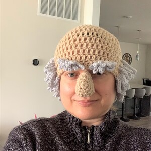 May include: A crocheted hat with a large, beige nose and gray hair. The hat is worn by a person with a neutral expression.