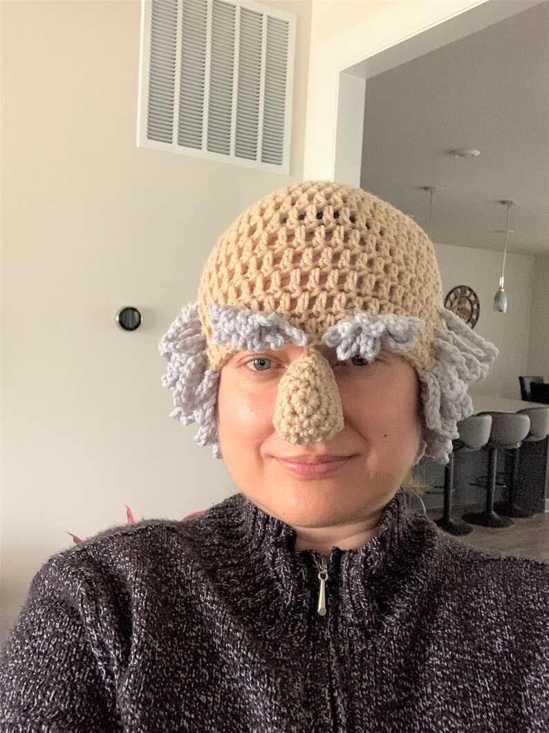 May include: A crocheted hat with a large, beige nose and gray hair. The hat is worn by a person with a neutral expression.
