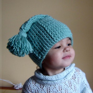 May include: A young child wearing a light blue knitted hat with a large pom-pom. The hat has a long, pointed top and is crocheted with a simple stitch pattern.