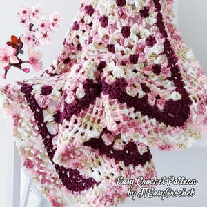 May include: A crocheted blanket in pink, white, and burgundy. The blanket has a floral design and a scalloped border. The text "Easy Crochet Pattern by JTEasyCrochet" is visible. The blanket is likely made with soft yarn.